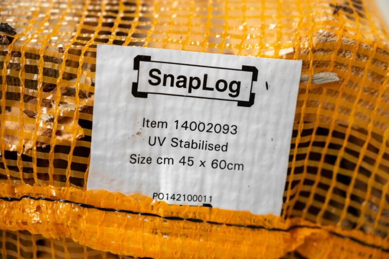 Closeup of a yellow bag of firewood labeled with item number UV-100000, indicating availability 24/7.